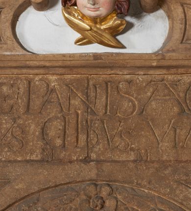 Zdjęcie nr 3: Sakramentarium w formie półkoliście zamkniętej niszy w kamiennym obramieniu o architektonicznych formach. Nisza ujęta arkadą  ustawioną na cokole dekorowanym rautami. Arkada wypełniona od frontu girlandą oraz trzema kwiatami. W przyłuczach trójkątne pola wypełnione kwiatami. Powyżej fryz z inskrypcją: „ECCE PANIS A(N)GELOR[UM] / FACTUS CIBUS VIATORU(M) / MM”. W zwieńczeniu wolutowo zawinięte odcinki przyczłóka. Pomiędzy nimi drewniana, polichromowana i złocona uskrzydlona główka anielska.