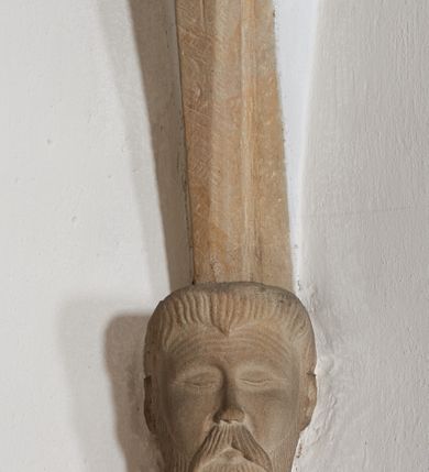 Zdjęcie nr 7: Komplet sześciu kamiennych konsol w formie męskich, brodatych głów, na które spływają żebra krzyżowego sklepienia w nawie głównej. Postać pierwsza o twarzy szerokiej, okolonej długim zarostem oraz włosami układającymi się w loki na czole. Oczy zamknięte, wydatny, prosty nos. Widoczne rurkowate bruzdy na czole. Postać druga o twarzy pociągłej, okolonej długim zarostem artykułowanym za pomocą rurkowatych fałd. Broda na końcu nieznacznie rozdwojona. Włosy proste ponad czołem tworzą trójkątne zakola. Oczy zamknięte, czoło pokryte bruzdami. Postać trzecia o trójkątnej twarzy z niskim, pokrytym bruzdami czołem i kręconych włosach. Oczy zamknięte. Postać czwarta o owalnej, pełnej twarzy, wystających kościach policzkowych. Zarost artykułowany za pomocą równoległych fałdek. Włosy układające się w loki ponad czołem. Oczy zamknięte. Postać piąta o szerokiej, płaskiej twarzy z gęstym zarostem. Długie, falowane wąsy, zamknięte oczy. Włosy ponad czołem układające się w loki. Postać szósta o pociągłej, podłużnej twarzy z zarostem. Długi, prosty nos, zamknięte oczy, niskie czoło ponad, którym bujne, proste włosy. Na przecięciach żeber dwa polichromowane zworniki z godłem herbu Drużyna oraz syglami „S(ebastian) L(ubomirski)&quot; umieszczonymi na czerwonym tle, pierwszy ośmioboczny, drugi w kształcie wieloliścia. W podniebiu arkady tęczowej wolutowa konsola ustawiona na kapitelu pilastra w podniebiu łuku tęczowego. U podstawy konsoli, na osi umieszczony rollwerkowy kartusz mieszczący godło herbu Drużyna oraz sygle „S(ebastian) L(ubomirski)”. 