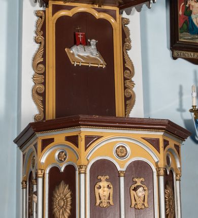 Zdjęcie nr 2: Ambona przyścienna, wisząca. Kosz na planie ośmioboku z cokołem i ośmiobocznym podwieszeniem. Cokół kosza dekorowany prostokątnymi płycinami. U spodu podwieszona kostka ujęta ze wszystkich stron wolutami oraz toczoną, profilowaną szyszką. Ścianki flankowane są kolumienkami podtrzymującymi półkoliste arkady w polach dekorowane biforiami. Biforia naprzemiennie ozdobione kwiatami słonecznika lub dzielone pośrodku kolumienką i zdobione dwiema uskrzydlonymi główkami aniołków w polach; nad biforiami w łuku arkady umieszczone są rozetki w medalionach. Zaplecek w formie wydłużonego, stojącego prostokąta z wpisaną w polu płyciną zamkniętą łukiem trójlistnym. Płycina ozdobiona jest płaskorzeźbionym przedstawieniem Baranka Eucharystycznego leżącego na księdze zamkniętej siedmioma pieczęciami. Po bokach zaplecek ujmują uszy z dekoracją roślinną. Baldachim na planie ośmioboku z cokołem dekorowanym płycinami, ornamentem roślinnym u góry i lambrekinem u spodu. Ścianki baldachimu zwężają się ku górze z aplikowanymi krzyżami i stanowią podstawę dla pełnej, czerwonej korony z krzyżem. U spodu baldachimu gołębica Ducha Świętego na tle promienistej glorii. Struktura polichromowana na bordowo, profilowania na żółto, detale i ornamentyka złocone i srebrzone.