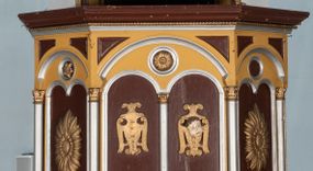 Zdjęcie nr 1: Ambona przyścienna, wisząca. Kosz na planie ośmioboku z cokołem i ośmiobocznym podwieszeniem. Cokół kosza dekorowany prostokątnymi płycinami. U spodu podwieszona kostka ujęta ze wszystkich stron wolutami oraz toczoną, profilowaną szyszką. Ścianki flankowane są kolumienkami podtrzymującymi półkoliste arkady w polach dekorowane biforiami. Biforia naprzemiennie ozdobione kwiatami słonecznika lub dzielone pośrodku kolumienką i zdobione dwiema uskrzydlonymi główkami aniołków w polach; nad biforiami w łuku arkady umieszczone są rozetki w medalionach. Zaplecek w formie wydłużonego, stojącego prostokąta z wpisaną w polu płyciną zamkniętą łukiem trójlistnym. Płycina ozdobiona jest płaskorzeźbionym przedstawieniem Baranka Eucharystycznego leżącego na księdze zamkniętej siedmioma pieczęciami. Po bokach zaplecek ujmują uszy z dekoracją roślinną. Baldachim na planie ośmioboku z cokołem dekorowanym płycinami, ornamentem roślinnym u góry i lambrekinem u spodu. Ścianki baldachimu zwężają się ku górze z aplikowanymi krzyżami i stanowią podstawę dla pełnej, czerwonej korony z krzyżem. U spodu baldachimu gołębica Ducha Świętego na tle promienistej glorii. Struktura polichromowana na bordowo, profilowania na żółto, detale i ornamentyka złocone i srebrzone.