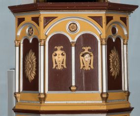 Zdjęcie nr 1: Ambona przyścienna, wisząca. Kosz na planie ośmioboku z cokołem i ośmiobocznym podwieszeniem. Cokół kosza dekorowany prostokątnymi płycinami. U spodu podwieszona kostka ujęta ze wszystkich stron wolutami oraz toczoną, profilowaną szyszką. Ścianki flankowane są kolumienkami podtrzymującymi półkoliste arkady w polach dekorowane biforiami. Biforia naprzemiennie ozdobione kwiatami słonecznika lub dzielone pośrodku kolumienką i zdobione dwiema uskrzydlonymi główkami aniołków w polach; nad biforiami w łuku arkady umieszczone są rozetki w medalionach. Zaplecek w formie wydłużonego, stojącego prostokąta z wpisaną w polu płyciną zamkniętą łukiem trójlistnym. Płycina ozdobiona jest płaskorzeźbionym przedstawieniem Baranka Eucharystycznego leżącego na księdze zamkniętej siedmioma pieczęciami. Po bokach zaplecek ujmują uszy z dekoracją roślinną. Baldachim na planie ośmioboku z cokołem dekorowanym płycinami, ornamentem roślinnym u góry i lambrekinem u spodu. Ścianki baldachimu zwężają się ku górze z aplikowanymi krzyżami i stanowią podstawę dla pełnej, czerwonej korony z krzyżem. U spodu baldachimu gołębica Ducha Świętego na tle promienistej glorii. Struktura polichromowana na bordowo, profilowania na żółto, detale i ornamentyka złocone i srebrzone.