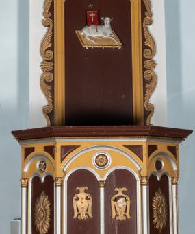 Zdjęcie nr 1: Ambona przyścienna, wisząca. Kosz na planie ośmioboku z cokołem i ośmiobocznym podwieszeniem. Cokół kosza dekorowany prostokątnymi płycinami. U spodu podwieszona kostka ujęta ze wszystkich stron wolutami oraz toczoną, profilowaną szyszką. Ścianki flankowane są kolumienkami podtrzymującymi półkoliste arkady w polach dekorowane biforiami. Biforia naprzemiennie ozdobione kwiatami słonecznika lub dzielone pośrodku kolumienką i zdobione dwiema uskrzydlonymi główkami aniołków w polach; nad biforiami w łuku arkady umieszczone są rozetki w medalionach. Zaplecek w formie wydłużonego, stojącego prostokąta z wpisaną w polu płyciną zamkniętą łukiem trójlistnym. Płycina ozdobiona jest płaskorzeźbionym przedstawieniem Baranka Eucharystycznego leżącego na księdze zamkniętej siedmioma pieczęciami. Po bokach zaplecek ujmują uszy z dekoracją roślinną. Baldachim na planie ośmioboku z cokołem dekorowanym płycinami, ornamentem roślinnym u góry i lambrekinem u spodu. Ścianki baldachimu zwężają się ku górze z aplikowanymi krzyżami i stanowią podstawę dla pełnej, czerwonej korony z krzyżem. U spodu baldachimu gołębica Ducha Świętego na tle promienistej glorii. Struktura polichromowana na bordowo, profilowania na żółto, detale i ornamentyka złocone i srebrzone.