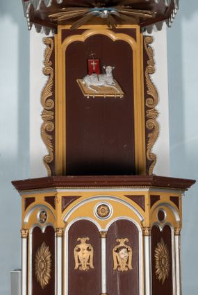 Zdjęcie nr 1: Ambona przyścienna, wisząca. Kosz na planie ośmioboku z cokołem i ośmiobocznym podwieszeniem. Cokół kosza dekorowany prostokątnymi płycinami. U spodu podwieszona kostka ujęta ze wszystkich stron wolutami oraz toczoną, profilowaną szyszką. Ścianki flankowane są kolumienkami podtrzymującymi półkoliste arkady w polach dekorowane biforiami. Biforia naprzemiennie ozdobione kwiatami słonecznika lub dzielone pośrodku kolumienką i zdobione dwiema uskrzydlonymi główkami aniołków w polach; nad biforiami w łuku arkady umieszczone są rozetki w medalionach. Zaplecek w formie wydłużonego, stojącego prostokąta z wpisaną w polu płyciną zamkniętą łukiem trójlistnym. Płycina ozdobiona jest płaskorzeźbionym przedstawieniem Baranka Eucharystycznego leżącego na księdze zamkniętej siedmioma pieczęciami. Po bokach zaplecek ujmują uszy z dekoracją roślinną. Baldachim na planie ośmioboku z cokołem dekorowanym płycinami, ornamentem roślinnym u góry i lambrekinem u spodu. Ścianki baldachimu zwężają się ku górze z aplikowanymi krzyżami i stanowią podstawę dla pełnej, czerwonej korony z krzyżem. U spodu baldachimu gołębica Ducha Świętego na tle promienistej glorii. Struktura polichromowana na bordowo, profilowania na żółto, detale i ornamentyka złocone i srebrzone.