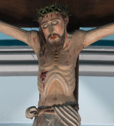 Zdjęcie nr 1: Krucyfiks z rzeźbą Chrystusa przybitą do krzyża trzema gwoździami. Figura zawieszona na szeroko rozłożonych, wyprężonych ramionach, w niewielkim zwisie. Sylwetka szczupła, o wyraźnie podkreślonej linii żeber i mostka. Głowa opada nieznacznie w kierunku prawego barku. Twarz podłużna, okolona krótką brodą, z zamkniętymi oczami; włosy długie, spływające na prawe ramię; na głowie korona cierniowa. Perizonium białe, przewiązane sznurem, odsłaniające prawe udo. Na ciele widoczne liczne strużki krwi spływające z ran. Krzyż prosty, gładki, ciemnobrązowy. Nad głową Chrystusa tabliczka w formie białej banderoli z napisem „IN / RI”.
