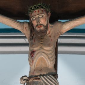 Zdjęcie nr 1: Krucyfiks z rzeźbą Chrystusa przybitą do krzyża trzema gwoździami. Figura zawieszona na szeroko rozłożonych, wyprężonych ramionach, w niewielkim zwisie. Sylwetka szczupła, o wyraźnie podkreślonej linii żeber i mostka. Głowa opada nieznacznie w kierunku prawego barku. Twarz podłużna, okolona krótką brodą, z zamkniętymi oczami; włosy długie, spływające na prawe ramię; na głowie korona cierniowa. Perizonium białe, przewiązane sznurem, odsłaniające prawe udo. Na ciele widoczne liczne strużki krwi spływające z ran. Krzyż prosty, gładki, ciemnobrązowy. Nad głową Chrystusa tabliczka w formie białej banderoli z napisem „IN / RI”.
