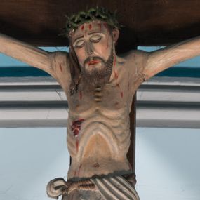 Zdjęcie nr 1: Krucyfiks z rzeźbą Chrystusa przybitą do krzyża trzema gwoździami. Figura zawieszona na szeroko rozłożonych, wyprężonych ramionach, w niewielkim zwisie. Sylwetka szczupła, o wyraźnie podkreślonej linii żeber i mostka. Głowa opada nieznacznie w kierunku prawego barku. Twarz podłużna, okolona krótką brodą, z zamkniętymi oczami; włosy długie, spływające na prawe ramię; na głowie korona cierniowa. Perizonium białe, przewiązane sznurem, odsłaniające prawe udo. Na ciele widoczne liczne strużki krwi spływające z ran. Krzyż prosty, gładki, ciemnobrązowy. Nad głową Chrystusa tabliczka w formie białej banderoli z napisem „IN / RI”.
