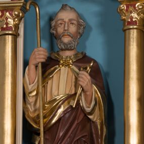 Zdjęcie nr 1: Rzeźba św. Piotra ustawiona na niskim prostopadłościennym cokole, o ściętych narożach. Święty Piotr ustawiony frontalnie, w prawej dłoni trzyma laskę pasterską, a w lewej dwa złote klucze. Twarz podłużna, ze zmarszczonym czołem, z długim nosem, dużymi oczami, okolona średniej długości brodą. Włosy siwe, kędzierzawe, z charakterystycznym kosmykiem nad czołem. Ubrany jest w długą beżową suknię z długimi rękawami oraz brązowy płaszcz ze złotą podszewką, zdobiony na brzegach złoconym pasem, spięty pod szyją dekoracyjną klamrą. Polichromia w odsłoniętych partiach ciała naturalistyczna, detale złocone.

