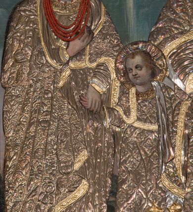 Zdjęcie nr 3: Obraz w kształcie stojącego prostokąta zamkniętego łukiem nadwieszonym, przedstawiająca Świętą Rodzinę. W centrum kompozycji mały Jezus trzyma za dłonie stojącego po prawej stronie obrazu św. Józefa oraz ukazaną po lewej stronie Matkę Boską. Postacie ukazane frontalnie, w postawie stojącej, Jezus z głową skierowaną w prawą stronę. Ponad nimi u góry nadlatująca gołębica Ducha Świętego. Maria przedstawiona jako młoda kobieta o pełnej, owalnej twarzy, okolonej długimi i falowanymi włosami. Prawą rękę wspiera na piersi, lewą trzyma za rękę Dzieciątko Jezus. Chrystus o pełnej, dziecięcej twarzy, okolonej jasnymi i krótkimi włosami. Święty Józef ukazany jako starszy mężczyzna z dłuższymi i siwymi włosami oraz krótką, gęstą brodą. Lewą rękę podtrzymuje rąbek szat, prawą trzyma za rękę Jezusa. Święty Józef oraz Matka Boska w koronach na głowie; wszystkie postacie w butach oraz długich i bogato drapowanych szatach przykrytych pozłacanymi sukienkami. W tle wypełniony roślinnością pejzaż oraz obłoki. Po lewej stronie św. Józefa gałązka białej lilii. Również na gołębicę Ducha Świętego nałożona srebrna sukienka. Kolorystyka chłodna, pociemniała. Rama drewniana, profilowana, złocona, zdobiona liśćmi stylizowanego akantu. 