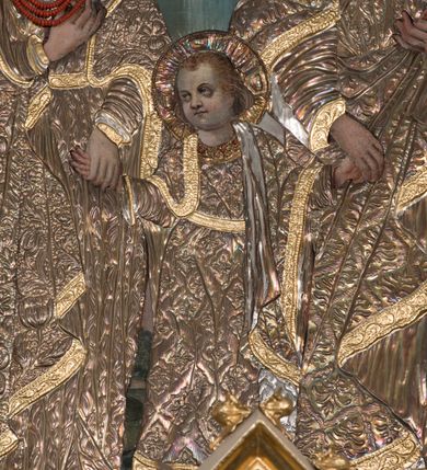 Zdjęcie nr 4: Obraz w kształcie stojącego prostokąta zamkniętego łukiem nadwieszonym, przedstawiająca Świętą Rodzinę. W centrum kompozycji mały Jezus trzyma za dłonie stojącego po prawej stronie obrazu św. Józefa oraz ukazaną po lewej stronie Matkę Boską. Postacie ukazane frontalnie, w postawie stojącej, Jezus z głową skierowaną w prawą stronę. Ponad nimi u góry nadlatująca gołębica Ducha Świętego. Maria przedstawiona jako młoda kobieta o pełnej, owalnej twarzy, okolonej długimi i falowanymi włosami. Prawą rękę wspiera na piersi, lewą trzyma za rękę Dzieciątko Jezus. Chrystus o pełnej, dziecięcej twarzy, okolonej jasnymi i krótkimi włosami. Święty Józef ukazany jako starszy mężczyzna z dłuższymi i siwymi włosami oraz krótką, gęstą brodą. Lewą rękę podtrzymuje rąbek szat, prawą trzyma za rękę Jezusa. Święty Józef oraz Matka Boska w koronach na głowie; wszystkie postacie w butach oraz długich i bogato drapowanych szatach przykrytych pozłacanymi sukienkami. W tle wypełniony roślinnością pejzaż oraz obłoki. Po lewej stronie św. Józefa gałązka białej lilii. Również na gołębicę Ducha Świętego nałożona srebrna sukienka. Kolorystyka chłodna, pociemniała. Rama drewniana, profilowana, złocona, zdobiona liśćmi stylizowanego akantu. 