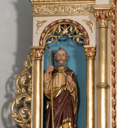 Zdjęcie nr 5: Ołtarz przyścienny, architektoniczny, trójosiowy z predellą i zwieńczeniem. Mensa prostopadłościenna, drewniana. Antepedium ujęte po bokach wysuniętymi przed lico lizenami z zakończonymi półokrągło płycinami. W polu o podziale architektonicznym – artykułowane złoconymi, kompozytowymi kolumienkami wspierającymi trójlistnie zakończone arkadki, w polu każdej dwie półkoliście zamknięte płyciny. Tabernakulum w formie prostopadłościennej skrzynki, flankowane ukośnie ustawionymi filarami wspierającymi profilowany gzyms. Drzwiczki wykonane z trybowanej, srebrzonej blachy; w polu środkowym z przedstawieniem głowy Chrystusa otoczonej gęstymi obłokami i promieniami, a dokoła ramą z wicią roślinną utworzoną z kiści winogron i kłosów zbóż. U spodu napis „TO JEST CIAŁO MOJE”. Na tabernakulum ustawiony jest tron wystawienia w formie prostopadłościennej aediculi, flankowany ukośnie ustawionymi kolumnami wspierającymi trójkątny szczyt. W polu prostokątna wnęka, zamknięta trójlistnie, o ściankach zdobionych motywem lilii heraldycznych. Po bokach tabernakulum ustawione dwie rzeźby klęczących aniołów.
Nastawa wsparta na wysokiej predelli, ujętej po bokach ażurowymi, akantowymi wolutami, pośrodku z dwiema lizenami wspierającymi podpory kondygnacji. W osiach predelli prostokątne płyciny, ozdobione w osiach bocznych rzeźbioną wicią roślinną. W polu środkowym kondygnacji głęboka, półkolista nisza mieszcząca obraz Świętej Rodziny, a na zasuwie obrazy Chrystusa Ukrzyżowanego i św. Józefa, obramowana płaskorzeźbioną ramą z liśćmi akantu; w podłuczu ażurowa akantowa dekoracja. Całość ujęta złoconymi kompozytowymi kolumnami, na których trzony w połowie wysokości nałożone są talerzyki. Kolumny dźwigają wysokie imposty oraz schodkowo przełamany gzyms. Ponad obrazem, w osi ołtarza znajdują się płaskorzeźbione główki anielskie o złoconych skrzydłach. Osie boczne o analogicznym układzie, arkady flankowane przez złocone kolumienki z figurami św. Piotra po lewej i św. Piotra po prawej stronie; zwieńczone pełnym belkowaniem – w partii fryzu symetryczny ornament o charakterze arabeskowym, gzyms dekorowany kimationem. Całość zwieńczona pinaklami zamkniętymi u góry złoconymi kwiatonami. Kondygnacja ujęta po bokach ażurowymi akantowymi uszami. Zwieńczenie flankowane złoconymi kolumienkami wspierającymi półkolistą arkadę oraz wyniesiony na osi szczyt ozdobiony pinaklami oraz w osi głównej krzyżem o trójlistnie zakończonych ramionach. W arkadzie ustawiona rzeźba Najświętszego Serca Pana Jezusa. Zwieńczenie ujęte po bokach ukształtowanymi w formę spływu uszami wypełnionymi płycinami z dekoracją akantową. Kolorystyka jasnobeżowa z elementami czerwieni, ornamenty i detale architektoniczne złocone.