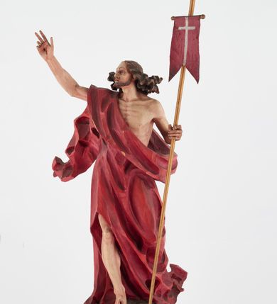Zdjęcie nr 10: Pełnoplastyczna figura Chrystusa Zmartwychwstałego ustawiona na prostopadłościennym postumencie o ściętych narożach. Chrystus ukazany w pozycji stojącej, w wykroku, z prawą nogą wysuniętą do przodu. Na stopie widoczna rana po gwoździach. Prawa ręka postaci uniesiona, lewa trzymająca drzewiec Chorągwi Zmartwychwstania zwieńczony krzyżem. Głowa Chrystusa lekko uniesiona, zwrócona w prawą stronę. Twarz pociągła o wyrazistych, ostrych rysach, okolona długimi, falowanymi, rozwianymi włosami i krótką, rozwidloną brodą. Chrystus ubrany w obszerny, czerwony płaszcz o głęboko rzeźbionych fałdach, z rozwianą połą przy prawym ramieniu. Figura polichromowana, drzewiec chorągwi złocony.