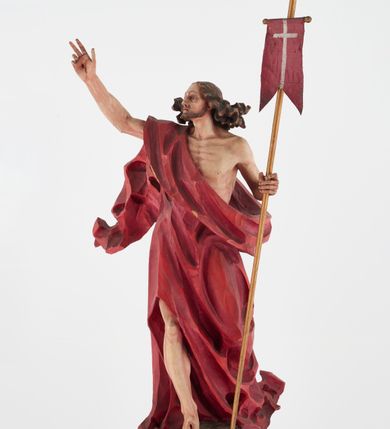 Zdjęcie nr 1: Pełnoplastyczna figura Chrystusa Zmartwychwstałego ustawiona na prostopadłościennym postumencie o ściętych narożach. Chrystus ukazany w pozycji stojącej, w wykroku, z prawą nogą wysuniętą do przodu. Na stopie widoczna rana po gwoździach. Prawa ręka postaci uniesiona, lewa trzymająca drzewiec Chorągwi Zmartwychwstania zwieńczony krzyżem. Głowa Chrystusa lekko uniesiona, zwrócona w prawą stronę. Twarz pociągła o wyrazistych, ostrych rysach, okolona długimi, falowanymi, rozwianymi włosami i krótką, rozwidloną brodą. Chrystus ubrany w obszerny, czerwony płaszcz o głęboko rzeźbionych fałdach, z rozwianą połą przy prawym ramieniu. Figura polichromowana, drzewiec chorągwi złocony.