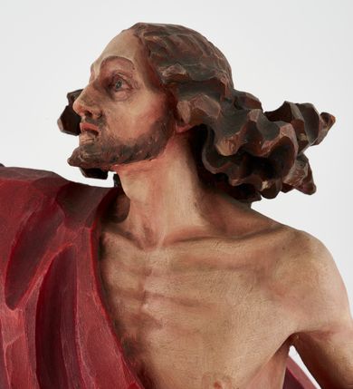 Zdjęcie nr 13: Pełnoplastyczna figura Chrystusa Zmartwychwstałego ustawiona na prostopadłościennym postumencie o ściętych narożach. Chrystus ukazany w pozycji stojącej, w wykroku, z prawą nogą wysuniętą do przodu. Na stopie widoczna rana po gwoździach. Prawa ręka postaci uniesiona, lewa trzymająca drzewiec Chorągwi Zmartwychwstania zwieńczony krzyżem. Głowa Chrystusa lekko uniesiona, zwrócona w prawą stronę. Twarz pociągła o wyrazistych, ostrych rysach, okolona długimi, falowanymi, rozwianymi włosami i krótką, rozwidloną brodą. Chrystus ubrany w obszerny, czerwony płaszcz o głęboko rzeźbionych fałdach, z rozwianą połą przy prawym ramieniu. Figura polichromowana, drzewiec chorągwi złocony.