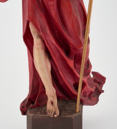 Zdjęcie nr 5: Pełnoplastyczna figura Chrystusa Zmartwychwstałego ustawiona na prostopadłościennym postumencie o ściętych narożach. Chrystus ukazany w pozycji stojącej, w wykroku, z prawą nogą wysuniętą do przodu. Na stopie widoczna rana po gwoździach. Prawa ręka postaci uniesiona, lewa trzymająca drzewiec Chorągwi Zmartwychwstania zwieńczony krzyżem. Głowa Chrystusa lekko uniesiona, zwrócona w prawą stronę. Twarz pociągła o wyrazistych, ostrych rysach, okolona długimi, falowanymi, rozwianymi włosami i krótką, rozwidloną brodą. Chrystus ubrany w obszerny, czerwony płaszcz o głęboko rzeźbionych fałdach, z rozwianą połą przy prawym ramieniu. Figura polichromowana, drzewiec chorągwi złocony.