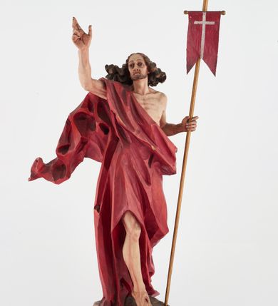 Zdjęcie nr 2: Pełnoplastyczna figura Chrystusa Zmartwychwstałego ustawiona na prostopadłościennym postumencie o ściętych narożach. Chrystus ukazany w pozycji stojącej, w wykroku, z prawą nogą wysuniętą do przodu. Na stopie widoczna rana po gwoździach. Prawa ręka postaci uniesiona, lewa trzymająca drzewiec Chorągwi Zmartwychwstania zwieńczony krzyżem. Głowa Chrystusa lekko uniesiona, zwrócona w prawą stronę. Twarz pociągła o wyrazistych, ostrych rysach, okolona długimi, falowanymi, rozwianymi włosami i krótką, rozwidloną brodą. Chrystus ubrany w obszerny, czerwony płaszcz o głęboko rzeźbionych fałdach, z rozwianą połą przy prawym ramieniu. Figura polichromowana, drzewiec chorągwi złocony.
