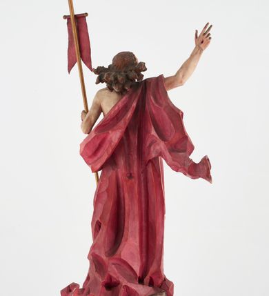 Zdjęcie nr 7: Pełnoplastyczna figura Chrystusa Zmartwychwstałego ustawiona na prostopadłościennym postumencie o ściętych narożach. Chrystus ukazany w pozycji stojącej, w wykroku, z prawą nogą wysuniętą do przodu. Na stopie widoczna rana po gwoździach. Prawa ręka postaci uniesiona, lewa trzymająca drzewiec Chorągwi Zmartwychwstania zwieńczony krzyżem. Głowa Chrystusa lekko uniesiona, zwrócona w prawą stronę. Twarz pociągła o wyrazistych, ostrych rysach, okolona długimi, falowanymi, rozwianymi włosami i krótką, rozwidloną brodą. Chrystus ubrany w obszerny, czerwony płaszcz o głęboko rzeźbionych fałdach, z rozwianą połą przy prawym ramieniu. Figura polichromowana, drzewiec chorągwi złocony.