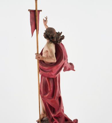 Zdjęcie nr 9: Pełnoplastyczna figura Chrystusa Zmartwychwstałego ustawiona na prostopadłościennym postumencie o ściętych narożach. Chrystus ukazany w pozycji stojącej, w wykroku, z prawą nogą wysuniętą do przodu. Na stopie widoczna rana po gwoździach. Prawa ręka postaci uniesiona, lewa trzymająca drzewiec Chorągwi Zmartwychwstania zwieńczony krzyżem. Głowa Chrystusa lekko uniesiona, zwrócona w prawą stronę. Twarz pociągła o wyrazistych, ostrych rysach, okolona długimi, falowanymi, rozwianymi włosami i krótką, rozwidloną brodą. Chrystus ubrany w obszerny, czerwony płaszcz o głęboko rzeźbionych fałdach, z rozwianą połą przy prawym ramieniu. Figura polichromowana, drzewiec chorągwi złocony.