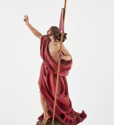 Zdjęcie nr 6: Pełnoplastyczna figura Chrystusa Zmartwychwstałego ustawiona na prostopadłościennym postumencie o ściętych narożach. Chrystus ukazany w pozycji stojącej, w wykroku, z prawą nogą wysuniętą do przodu. Na stopie widoczna rana po gwoździach. Prawa ręka postaci uniesiona, lewa trzymająca drzewiec Chorągwi Zmartwychwstania zwieńczony krzyżem. Głowa Chrystusa lekko uniesiona, zwrócona w prawą stronę. Twarz pociągła o wyrazistych, ostrych rysach, okolona długimi, falowanymi, rozwianymi włosami i krótką, rozwidloną brodą. Chrystus ubrany w obszerny, czerwony płaszcz o głęboko rzeźbionych fałdach, z rozwianą połą przy prawym ramieniu. Figura polichromowana, drzewiec chorągwi złocony.