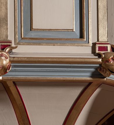 Zdjęcie nr 7: Ambona przyścienna, wisząca, składająca się z kosza, zaplecka i baldachimu, z dostępem od lewej strony w postaci jednobiegowych schodów z balustradą. Kosz na planie sześcioboku, o ściankach prostych, dekorowanych płycinami w kształcie stojących prostokątów zakończonych łukiem półkolistym. Płyciny rozdzilają pilastry o kompozytowych głowicach, posadowione na konsolach utworzonych z główek atrybutów czterech ewangelistów i podtrzymujących arkadki dekorowane zygzakiem. Gzyms wieńczący kosz zdobiony dwoma rzędami ornamentu geometrycznego. Podstawa kosza stożkowa, z wypełniającymi płaszczyzny płycinami, przechodząca w wieloboczny filar, którego głowica utworzona została z liści akantu. Zaplecek w kształcie prostokąta z prostokątną płyciną zamkniętą łukiem półkolistym. Baldachim powtarzający plan kosza, ujęty profilowanym gzymsem, z podwieszeniem z arkadek i zwieńczony na załamaniach liśćmi. Na jego szczycie pełnoplastyczna figura Chrystusa Pantokratora ustawiona na stożkowej podstawie w formie fragmentu filara. Chrystus został ukazany w całej postaci, z prawą ręką uniesioną w geście błogosławieństwa, lewą trzyma glob. Ma owalną twarz okoloną długimi włosami, które spływają mu na ramiona i plecy; otaczający głowę nimb przybiera formę trzech wiązek promieni. Jest ubrany w czerwoną suknienkę i złoty płaszcz; szaty łamią się ostrymi fałdami. Balustrada z poręczą z tralkami spiętymi ażurowymi arkadkami. Kolorystyka pastelowa, jasna. Struktura w kolorze kremowym, płyciny jasnoszare, fryzy bordowe, detal architektoniczny i ornamenty pozłacane.
