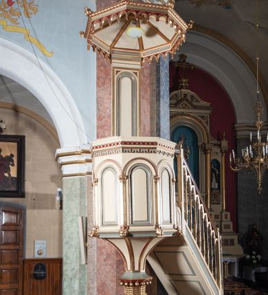 Zdjęcie nr 1: Ambona przyścienna, wisząca, składająca się z kosza, zaplecka i baldachimu, z dostępem od lewej strony w postaci jednobiegowych schodów z balustradą. Kosz na planie sześcioboku, o ściankach prostych, dekorowanych płycinami w kształcie stojących prostokątów zakończonych łukiem półkolistym. Płyciny rozdzilają pilastry o kompozytowych głowicach, posadowione na konsolach utworzonych z główek atrybutów czterech ewangelistów i podtrzymujących arkadki dekorowane zygzakiem. Gzyms wieńczący kosz zdobiony dwoma rzędami ornamentu geometrycznego. Podstawa kosza stożkowa, z wypełniającymi płaszczyzny płycinami, przechodząca w wieloboczny filar, którego głowica utworzona została z liści akantu. Zaplecek w kształcie prostokąta z prostokątną płyciną zamkniętą łukiem półkolistym. Baldachim powtarzający plan kosza, ujęty profilowanym gzymsem, z podwieszeniem z arkadek i zwieńczony na załamaniach liśćmi. Na jego szczycie pełnoplastyczna figura Chrystusa Pantokratora ustawiona na stożkowej podstawie w formie fragmentu filara. Chrystus został ukazany w całej postaci, z prawą ręką uniesioną w geście błogosławieństwa, lewą trzyma glob. Ma owalną twarz okoloną długimi włosami, które spływają mu na ramiona i plecy; otaczający głowę nimb przybiera formę trzech wiązek promieni. Jest ubrany w czerwoną suknienkę i złoty płaszcz; szaty łamią się ostrymi fałdami. Balustrada z poręczą z tralkami spiętymi ażurowymi arkadkami. Kolorystyka pastelowa, jasna. Struktura w kolorze kremowym, płyciny jasnoszare, fryzy bordowe, detal architektoniczny i ornamenty pozłacane.
