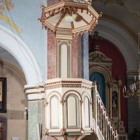 Zdjęcie nr 1: Ambona przyścienna, wisząca, składająca się z kosza, zaplecka i baldachimu, z dostępem od lewej strony w postaci jednobiegowych schodów z balustradą. Kosz na planie sześcioboku, o ściankach prostych, dekorowanych płycinami w kształcie stojących prostokątów zakończonych łukiem półkolistym. Płyciny rozdzilają pilastry o kompozytowych głowicach, posadowione na konsolach utworzonych z główek atrybutów czterech ewangelistów i podtrzymujących arkadki dekorowane zygzakiem. Gzyms wieńczący kosz zdobiony dwoma rzędami ornamentu geometrycznego. Podstawa kosza stożkowa, z wypełniającymi płaszczyzny płycinami, przechodząca w wieloboczny filar, którego głowica utworzona została z liści akantu. Zaplecek w kształcie prostokąta z prostokątną płyciną zamkniętą łukiem półkolistym. Baldachim powtarzający plan kosza, ujęty profilowanym gzymsem, z podwieszeniem z arkadek i zwieńczony na załamaniach liśćmi. Na jego szczycie pełnoplastyczna figura Chrystusa Pantokratora ustawiona na stożkowej podstawie w formie fragmentu filara. Chrystus został ukazany w całej postaci, z prawą ręką uniesioną w geście błogosławieństwa, lewą trzyma glob. Ma owalną twarz okoloną długimi włosami, które spływają mu na ramiona i plecy; otaczający głowę nimb przybiera formę trzech wiązek promieni. Jest ubrany w czerwoną suknienkę i złoty płaszcz; szaty łamią się ostrymi fałdami. Balustrada z poręczą z tralkami spiętymi ażurowymi arkadkami. Kolorystyka pastelowa, jasna. Struktura w kolorze kremowym, płyciny jasnoszare, fryzy bordowe, detal architektoniczny i ornamenty pozłacane.
