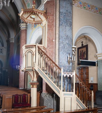 Zdjęcie nr 2: Ambona przyścienna, wisząca, składająca się z kosza, zaplecka i baldachimu, z dostępem od lewej strony w postaci jednobiegowych schodów z balustradą. Kosz na planie sześcioboku, o ściankach prostych, dekorowanych płycinami w kształcie stojących prostokątów zakończonych łukiem półkolistym. Płyciny rozdzilają pilastry o kompozytowych głowicach, posadowione na konsolach utworzonych z główek atrybutów czterech ewangelistów i podtrzymujących arkadki dekorowane zygzakiem. Gzyms wieńczący kosz zdobiony dwoma rzędami ornamentu geometrycznego. Podstawa kosza stożkowa, z wypełniającymi płaszczyzny płycinami, przechodząca w wieloboczny filar, którego głowica utworzona została z liści akantu. Zaplecek w kształcie prostokąta z prostokątną płyciną zamkniętą łukiem półkolistym. Baldachim powtarzający plan kosza, ujęty profilowanym gzymsem, z podwieszeniem z arkadek i zwieńczony na załamaniach liśćmi. Na jego szczycie pełnoplastyczna figura Chrystusa Pantokratora ustawiona na stożkowej podstawie w formie fragmentu filara. Chrystus został ukazany w całej postaci, z prawą ręką uniesioną w geście błogosławieństwa, lewą trzyma glob. Ma owalną twarz okoloną długimi włosami, które spływają mu na ramiona i plecy; otaczający głowę nimb przybiera formę trzech wiązek promieni. Jest ubrany w czerwoną suknienkę i złoty płaszcz; szaty łamią się ostrymi fałdami. Balustrada z poręczą z tralkami spiętymi ażurowymi arkadkami. Kolorystyka pastelowa, jasna. Struktura w kolorze kremowym, płyciny jasnoszare, fryzy bordowe, detal architektoniczny i ornamenty pozłacane.
