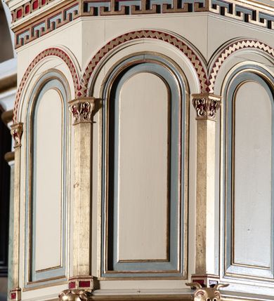 Zdjęcie nr 6: Ambona przyścienna, wisząca, składająca się z kosza, zaplecka i baldachimu, z dostępem od lewej strony w postaci jednobiegowych schodów z balustradą. Kosz na planie sześcioboku, o ściankach prostych, dekorowanych płycinami w kształcie stojących prostokątów zakończonych łukiem półkolistym. Płyciny rozdzilają pilastry o kompozytowych głowicach, posadowione na konsolach utworzonych z główek atrybutów czterech ewangelistów i podtrzymujących arkadki dekorowane zygzakiem. Gzyms wieńczący kosz zdobiony dwoma rzędami ornamentu geometrycznego. Podstawa kosza stożkowa, z wypełniającymi płaszczyzny płycinami, przechodząca w wieloboczny filar, którego głowica utworzona została z liści akantu. Zaplecek w kształcie prostokąta z prostokątną płyciną zamkniętą łukiem półkolistym. Baldachim powtarzający plan kosza, ujęty profilowanym gzymsem, z podwieszeniem z arkadek i zwieńczony na załamaniach liśćmi. Na jego szczycie pełnoplastyczna figura Chrystusa Pantokratora ustawiona na stożkowej podstawie w formie fragmentu filara. Chrystus został ukazany w całej postaci, z prawą ręką uniesioną w geście błogosławieństwa, lewą trzyma glob. Ma owalną twarz okoloną długimi włosami, które spływają mu na ramiona i plecy; otaczający głowę nimb przybiera formę trzech wiązek promieni. Jest ubrany w czerwoną suknienkę i złoty płaszcz; szaty łamią się ostrymi fałdami. Balustrada z poręczą z tralkami spiętymi ażurowymi arkadkami. Kolorystyka pastelowa, jasna. Struktura w kolorze kremowym, płyciny jasnoszare, fryzy bordowe, detal architektoniczny i ornamenty pozłacane.
