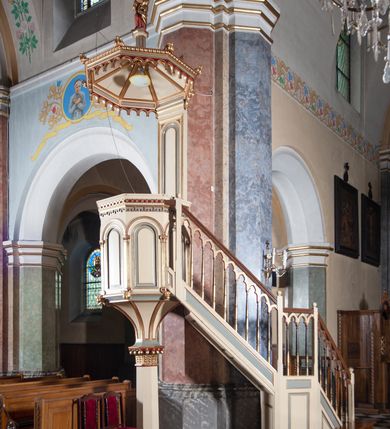 Zdjęcie nr 3: Ambona przyścienna, wisząca, składająca się z kosza, zaplecka i baldachimu, z dostępem od lewej strony w postaci jednobiegowych schodów z balustradą. Kosz na planie sześcioboku, o ściankach prostych, dekorowanych płycinami w kształcie stojących prostokątów zakończonych łukiem półkolistym. Płyciny rozdzilają pilastry o kompozytowych głowicach, posadowione na konsolach utworzonych z główek atrybutów czterech ewangelistów i podtrzymujących arkadki dekorowane zygzakiem. Gzyms wieńczący kosz zdobiony dwoma rzędami ornamentu geometrycznego. Podstawa kosza stożkowa, z wypełniającymi płaszczyzny płycinami, przechodząca w wieloboczny filar, którego głowica utworzona została z liści akantu. Zaplecek w kształcie prostokąta z prostokątną płyciną zamkniętą łukiem półkolistym. Baldachim powtarzający plan kosza, ujęty profilowanym gzymsem, z podwieszeniem z arkadek i zwieńczony na załamaniach liśćmi. Na jego szczycie pełnoplastyczna figura Chrystusa Pantokratora ustawiona na stożkowej podstawie w formie fragmentu filara. Chrystus został ukazany w całej postaci, z prawą ręką uniesioną w geście błogosławieństwa, lewą trzyma glob. Ma owalną twarz okoloną długimi włosami, które spływają mu na ramiona i plecy; otaczający głowę nimb przybiera formę trzech wiązek promieni. Jest ubrany w czerwoną suknienkę i złoty płaszcz; szaty łamią się ostrymi fałdami. Balustrada z poręczą z tralkami spiętymi ażurowymi arkadkami. Kolorystyka pastelowa, jasna. Struktura w kolorze kremowym, płyciny jasnoszare, fryzy bordowe, detal architektoniczny i ornamenty pozłacane.
