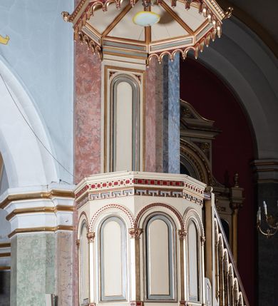 Zdjęcie nr 4: Ambona przyścienna, wisząca, składająca się z kosza, zaplecka i baldachimu, z dostępem od lewej strony w postaci jednobiegowych schodów z balustradą. Kosz na planie sześcioboku, o ściankach prostych, dekorowanych płycinami w kształcie stojących prostokątów zakończonych łukiem półkolistym. Płyciny rozdzilają pilastry o kompozytowych głowicach, posadowione na konsolach utworzonych z główek atrybutów czterech ewangelistów i podtrzymujących arkadki dekorowane zygzakiem. Gzyms wieńczący kosz zdobiony dwoma rzędami ornamentu geometrycznego. Podstawa kosza stożkowa, z wypełniającymi płaszczyzny płycinami, przechodząca w wieloboczny filar, którego głowica utworzona została z liści akantu. Zaplecek w kształcie prostokąta z prostokątną płyciną zamkniętą łukiem półkolistym. Baldachim powtarzający plan kosza, ujęty profilowanym gzymsem, z podwieszeniem z arkadek i zwieńczony na załamaniach liśćmi. Na jego szczycie pełnoplastyczna figura Chrystusa Pantokratora ustawiona na stożkowej podstawie w formie fragmentu filara. Chrystus został ukazany w całej postaci, z prawą ręką uniesioną w geście błogosławieństwa, lewą trzyma glob. Ma owalną twarz okoloną długimi włosami, które spływają mu na ramiona i plecy; otaczający głowę nimb przybiera formę trzech wiązek promieni. Jest ubrany w czerwoną suknienkę i złoty płaszcz; szaty łamią się ostrymi fałdami. Balustrada z poręczą z tralkami spiętymi ażurowymi arkadkami. Kolorystyka pastelowa, jasna. Struktura w kolorze kremowym, płyciny jasnoszare, fryzy bordowe, detal architektoniczny i ornamenty pozłacane.
