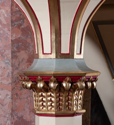 Zdjęcie nr 8: Ambona przyścienna, wisząca, składająca się z kosza, zaplecka i baldachimu, z dostępem od lewej strony w postaci jednobiegowych schodów z balustradą. Kosz na planie sześcioboku, o ściankach prostych, dekorowanych płycinami w kształcie stojących prostokątów zakończonych łukiem półkolistym. Płyciny rozdzilają pilastry o kompozytowych głowicach, posadowione na konsolach utworzonych z główek atrybutów czterech ewangelistów i podtrzymujących arkadki dekorowane zygzakiem. Gzyms wieńczący kosz zdobiony dwoma rzędami ornamentu geometrycznego. Podstawa kosza stożkowa, z wypełniającymi płaszczyzny płycinami, przechodząca w wieloboczny filar, którego głowica utworzona została z liści akantu. Zaplecek w kształcie prostokąta z prostokątną płyciną zamkniętą łukiem półkolistym. Baldachim powtarzający plan kosza, ujęty profilowanym gzymsem, z podwieszeniem z arkadek i zwieńczony na załamaniach liśćmi. Na jego szczycie pełnoplastyczna figura Chrystusa Pantokratora ustawiona na stożkowej podstawie w formie fragmentu filara. Chrystus został ukazany w całej postaci, z prawą ręką uniesioną w geście błogosławieństwa, lewą trzyma glob. Ma owalną twarz okoloną długimi włosami, które spływają mu na ramiona i plecy; otaczający głowę nimb przybiera formę trzech wiązek promieni. Jest ubrany w czerwoną suknienkę i złoty płaszcz; szaty łamią się ostrymi fałdami. Balustrada z poręczą z tralkami spiętymi ażurowymi arkadkami. Kolorystyka pastelowa, jasna. Struktura w kolorze kremowym, płyciny jasnoszare, fryzy bordowe, detal architektoniczny i ornamenty pozłacane.
