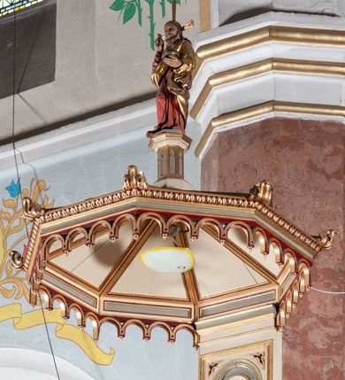 Zdjęcie nr 5: Ambona przyścienna, wisząca, składająca się z kosza, zaplecka i baldachimu, z dostępem od lewej strony w postaci jednobiegowych schodów z balustradą. Kosz na planie sześcioboku, o ściankach prostych, dekorowanych płycinami w kształcie stojących prostokątów zakończonych łukiem półkolistym. Płyciny rozdzilają pilastry o kompozytowych głowicach, posadowione na konsolach utworzonych z główek atrybutów czterech ewangelistów i podtrzymujących arkadki dekorowane zygzakiem. Gzyms wieńczący kosz zdobiony dwoma rzędami ornamentu geometrycznego. Podstawa kosza stożkowa, z wypełniającymi płaszczyzny płycinami, przechodząca w wieloboczny filar, którego głowica utworzona została z liści akantu. Zaplecek w kształcie prostokąta z prostokątną płyciną zamkniętą łukiem półkolistym. Baldachim powtarzający plan kosza, ujęty profilowanym gzymsem, z podwieszeniem z arkadek i zwieńczony na załamaniach liśćmi. Na jego szczycie pełnoplastyczna figura Chrystusa Pantokratora ustawiona na stożkowej podstawie w formie fragmentu filara. Chrystus został ukazany w całej postaci, z prawą ręką uniesioną w geście błogosławieństwa, lewą trzyma glob. Ma owalną twarz okoloną długimi włosami, które spływają mu na ramiona i plecy; otaczający głowę nimb przybiera formę trzech wiązek promieni. Jest ubrany w czerwoną suknienkę i złoty płaszcz; szaty łamią się ostrymi fałdami. Balustrada z poręczą z tralkami spiętymi ażurowymi arkadkami. Kolorystyka pastelowa, jasna. Struktura w kolorze kremowym, płyciny jasnoszare, fryzy bordowe, detal architektoniczny i ornamenty pozłacane.
