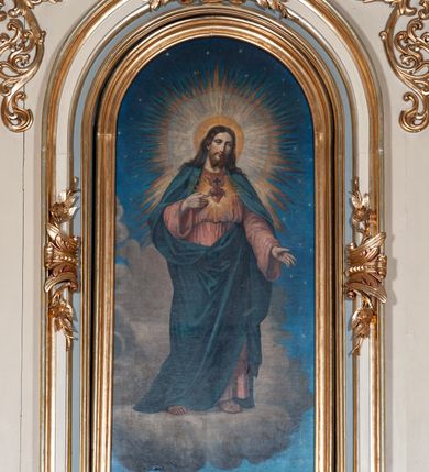 Zdjęcie nr 2: Obraz w kształcie stojącego prostokąta z przedstawieniem Najświętszego Serca Jezusa ujęty złotą, profilowaną ramą. W centrum kompozycji Chrystus ukazany w całej postaci. Prawą ręką wskazuje na gorejące, koronowane cierniem serce umieszczone na jego piersi, lewą ma opuszczoną, głowę przechyla na lewe ramię, patrzy na widza. Ma owalną twarz o smutnym wyrazie, brązowe oczy, wąski nos i niewielkie usta, na policzkach delikatny rumieniec. Długie, brązowe włosy spływają mu na ramiona i plecy, twarz okala rozdzielona w dwa pukle broda i wąsy. Wokół głowy szeroki, promienisty nimb w różnych odcieniach żółtego koloru. Chrystus jest ubrany w długą, jasnoczerwoną suknię przepasaną sznurem, a na nią ma zarzucony ciemnoniebieski płaszcz, który zakrywa nogi, ramiona i jest zaczepiony o pasek przy lewym biodrze. Zbawiciel stoi na i pośród chmur, w tle gwieździste niebo. Kolorystyka obrazu jasna, pastelowa.   
