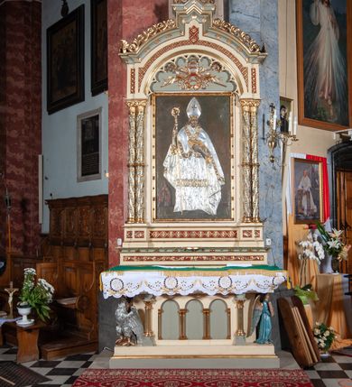 Zdjęcie nr 2: Ołtarz architektoniczny, jednokondygnacyjny, jednoosiowy, w formie aediculi, na podwójnym cokole oddzielonym linią gzymsu. Antepedium w formie leżącego prostokąta z trzema arkadami podtrzymywanymi przez kolumny toskańskie. Całość ujęta w dwie grube kolumny o kapitelach kompozytowych, stojące na cokołach. W narożach figury czterech istot apokaliptycznych: lwa, orła, anioła i wołu.  Pole środkowe nastawy flankowane jest dwiema parami kolumn na cokołach, o kapitelach kompozytowych. Wewnętrzna para kolumn została wysunięta nieco przed lico ołtarza i podtrzymuje półokrągłą arkadę. Całość zwieńczona gzymsem w formie łuku nadwieszonego, który w partii środkowej wyłamuje się, przybierając kształt prostokąta zwieńczonego trójkątnie. Cokoły dekorowane płycinami z ornamentem roślinnym, geometryczno-roślinnym i rozetami. Trzony kolumn kanelowane spiralnie, w połowie ujęte opaską, dekorowane guzami. Archiwolta arkady profilowana, ozdobiona maswerkiem. W polu środkowym obraz św. Mikołaja w profilowanej ramie z elementami geometrycznymi i liśćmi. W polu tympanonu płaskorzeźba z insygniami papieskimi – infułą, pastorałem, krzyżem patriarszym i kluczami. Powierzchnia ponad arkadą dekorowana kratką regencyjną z rozetami. Gzyms wieńczący ozdobiony rozetami i liśćmi oraz ażurowym akantem. Całość zwieńczona krzyżem. 
Struktura polichromowana na kolor kremowy, płyciny niebieskie i ciemnoczerwone, detale i ornamenty złocone i srebrzone. 
