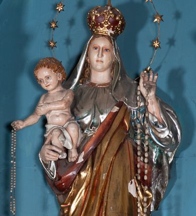 Zdjęcie nr 1: Figura Matki Boskiej z Dzieciątkiem w typie Różańcowej. Maria ujęta frontalnie, prawą ręką podtrzymuje Dzieciątko, na lewej ma zawieszony różaniec. Spogląda przed siebie, ma małą głowę osadzoną na szerokiej szyi, niewielkie, niebieskie oczy i wąski nos; włosy przykrywa welon. Jest ubrana w srebrną suknię lamowaną pod szyją złotem oraz złoty płaszcz podbity czerwienią przechodzący przez lewe ramię. Ma głowie ma koronę zamknietą zwieńczona krzyżykiem, wokół głowy nimb z 12 gwiazd. Stoi na połowie kuli ziemskiej, pod lewą stopą ma półksiężyc, pod prawą węża, po półkuli w dół toczy się jabłko. Dzieciątko spogląda przed siebie, w prawej ręce trzyma różaniec, lewą wyciąga przed siebie; biodra ma przepasane białą szarfą. Ma małą, okrągłą głowę, okoloną jasnymi, kręconymi włosami. 