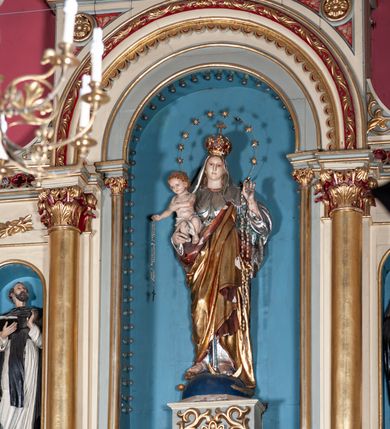 Zdjęcie nr 3: Figura Matki Boskiej z Dzieciątkiem w typie Różańcowej. Maria ujęta frontalnie, prawą ręką podtrzymuje Dzieciątko, na lewej ma zawieszony różaniec. Spogląda przed siebie, ma małą głowę osadzoną na szerokiej szyi, niewielkie, niebieskie oczy i wąski nos; włosy przykrywa welon. Jest ubrana w srebrną suknię lamowaną pod szyją złotem oraz złoty płaszcz podbity czerwienią przechodzący przez lewe ramię. Ma głowie ma koronę zamknietą zwieńczona krzyżykiem, wokół głowy nimb z 12 gwiazd. Stoi na połowie kuli ziemskiej, pod lewą stopą ma półksiężyc, pod prawą węża, po półkuli w dół toczy się jabłko. Dzieciątko spogląda przed siebie, w prawej ręce trzyma różaniec, lewą wyciąga przed siebie; biodra ma przepasane białą szarfą. Ma małą, okrągłą głowę, okoloną jasnymi, kręconymi włosami. 