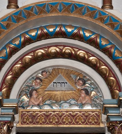 Zdjęcie nr 3: Ołtarz architektoniczny, jednokondygnacyjny, trójosiowy ze zwieńczeniem i bramkami, ustawiony na cokole. Mensa marmurowa z portatylem. Antepedium w formie wydłużonego, leżącego prostokąta. Ujęte w profilowaną ramę, podzielone na trzy części. Partia środkowa kwadratowa, w niej medalion z płaskorzeźbionym przedstawieniem gorejącego serca na tle skrzyżowanego krzyża i kotwicy. W partiach bocznych płaskorzeźbiony motyw maswerku. Na mensie prostopadłościenne tabernakulum z drzwiczkami dekorowanymi motywem silnie stylizowanych winnych gron, ustawione na cokole przerywanym trzema plakietami roślinnymi. Tabernakulum z dwóch stron podtrzymują stylizowane anioły. Pole środkowe nastawy ołtarza w formie uskokowego portalu zamkniętego półokrągłym łukiem z tympanonem. Dwie wewnętrzne archiwolty podtrzymywane przez półkolumny z kapitelami kompozytowymi, zewnętrzna przez pilastry. Portal flankują dwie nisze, zwieńczone łukiem nadwieszonym. Pierwszą kondygnację zamyka pas fryzu. W polu zwieńczenia ponad łukiem portalu rozchodzi się stopniowo pięć par kolumn o kostkowych kapitelach, które w partii wieńczącej podtrzymują profilowany łuk nadwieszony. Zwieńczenie zamknięte trójkątną linią belkowania, z pasem stopniowo spływających wolut, zakończone krzyżem. Cokół dekorowany płycinami z perełkowaniem, zakończony gzymsem z motywem wolich oczu. Poszczególne elementy niszy dekorowane różnorodnym ornamentem o charakterze geometryczno-roślinnym. W polu środkowym obraz Matki Boskiej z Dzieciątkiem w srebrnej sukience (Matki Boskiej Lisieckiej). W tympanonie płaskorzeźbione przedstawienie imienia Boga ujętego w trójkąt w glorii promienistej, w otoczeniu putt pośród obłoków. Nisze flankujące portal zwieńczone są płaskorzeźbionymi przedstawieniami centralnych świątyń kopułowych. W niszach płaskorzeźby św. Piotra po lewej i św. Pawła po prawej stronie. Fryz zamykający kondygnację z motywem palmet, w miejscu przecięcia z opaską portalu – uskrzydlone główki anielskie. W partii zwieńczenia, w przestrzeniach ponad kolumnami rozety. Ostatnia para kolumn ujęta w pionowe pasy imitujące zwinięty sznur. Łuk nadwieszony dekorowany ornamentem roślinnym. Cały ołtarz ujęty w pasy z dekoracją roślinną i motywem sznura. Wieńczące belkowanie z dekoracją geometryczną i rozetami, ujęte w woluty z mięsistego akantu. Podstawę dla krzyża tworzy akant z motywem palmety i perełkowania. Ołtarz flankują dwie bramki, na których stoją figury św. Michała Archanioła po lewej i św. Józefa po prawej stronie. 
Struktura w kolorze naturalnego kamienia – kremowa, profile w odcieniach niebieskiego, detale i ornamenty złocone. 
