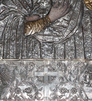 Zdjęcie nr 5: Obraz w formie stojącego prostokąta. Matka Boska zwrócona w trzech czwartych w lewo. Obiema rękami przytula Dzieciątko. Ma delikatną, owalną twarz o wąskich brwiach, niewielkich, ciemnych oczach, wąskim nosie i małych ustach, włosy skrywa maforion. Dzieciątko ukazane zostało niemal profilem, a jego głowa odwrócona jest w stronę widza. Prawą ręką dotyka twarzy Marii, lewą  kładzie jej na dekolcie, jedną stopę opiera na jej ręce. Ma okrągłą twarz o rysach ukazujących podobieństwo do Matki. Sukienki postaci oraz tło zostały wykonane z trybowanej, srebrnej blachy pozłoconej w niektórych partiach. Maria jest ubrana w złotą tunikę, z której widać jedynie fragmenty rękawów i dekoltu, na to ma nałożony maforion dekorowany w drobny ornament roślinny. Dzieciątko ma na sobie sukienkę o podobnym wzorze, podbitą złotem. Oboje mają na głowach korony zamknięte dekorowane kameryzacją. Korona Dzieciątka jest złota, a srebrną koronę Marii podtrzymują dwa, złocone anioły w długich sukniach. Tło trybowane w większy wzór o motywach rozwiniętych kwiatów. Od góry i od dołu obraz powiększono o metalowe fryzy. W górnej partii obrazu dodano pas ze złoconą dekoracją roślinną, w dolnej zaś z przedstawieniem krzyża i dwóch kartuszy herbowych: Orła i Pogoni, w otoczeniu dekoracji o motywie roślinnym. Obraz w wypukłej ramie o dekoracji roślinnej. 