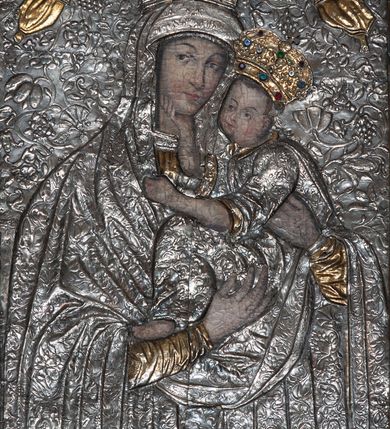 Zdjęcie nr 4: Obraz w formie stojącego prostokąta. Matka Boska zwrócona w trzech czwartych w lewo. Obiema rękami przytula Dzieciątko. Ma delikatną, owalną twarz o wąskich brwiach, niewielkich, ciemnych oczach, wąskim nosie i małych ustach, włosy skrywa maforion. Dzieciątko ukazane zostało niemal profilem, a jego głowa odwrócona jest w stronę widza. Prawą ręką dotyka twarzy Marii, lewą  kładzie jej na dekolcie, jedną stopę opiera na jej ręce. Ma okrągłą twarz o rysach ukazujących podobieństwo do Matki. Sukienki postaci oraz tło zostały wykonane z trybowanej, srebrnej blachy pozłoconej w niektórych partiach. Maria jest ubrana w złotą tunikę, z której widać jedynie fragmenty rękawów i dekoltu, na to ma nałożony maforion dekorowany w drobny ornament roślinny. Dzieciątko ma na sobie sukienkę o podobnym wzorze, podbitą złotem. Oboje mają na głowach korony zamknięte dekorowane kameryzacją. Korona Dzieciątka jest złota, a srebrną koronę Marii podtrzymują dwa, złocone anioły w długich sukniach. Tło trybowane w większy wzór o motywach rozwiniętych kwiatów. Od góry i od dołu obraz powiększono o metalowe fryzy. W górnej partii obrazu dodano pas ze złoconą dekoracją roślinną, w dolnej zaś z przedstawieniem krzyża i dwóch kartuszy herbowych: Orła i Pogoni, w otoczeniu dekoracji o motywie roślinnym. Obraz w wypukłej ramie o dekoracji roślinnej. 