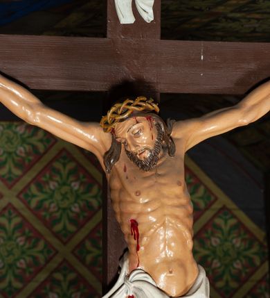 Zdjęcie nr 2: Trójfigurowa grupa Ukrzyżowania, rzeźby pełne. W środku znajduje się krucyfiks z postacią umarłego Chrystusa przybitego trzema gwoździami do krzyża. Chrystus w lekkim zwisie, o szeroko rozłożonych ramionach, bezwładny, głowa opuszczona na prawe ramię. Twarz szczupła, owalna, o prostym nosie, dużych oczach i na wpół otwartych ustach, okolona zarostem. Włosy w postaci delikatnych fal opadają na plecy i prawe ramię. Na głowie korona cierniowa. Ciało o wyraźnie zarysowanej anatomii. Perizonium w formie tkaniny przewiązanej na boku i odsłaniającej prawe biodro, z dłuższym brzegiem zwisającym po prawej stronie. Draperia gęsto fałdowana, o miękkich załamaniach materiału. Na szczycie pionowej belki titulus w formie banderoli z napisem „IN/RI”. Figura Matki Boskiej ustawiona po lewej stronie, zwrócona lekko w stronę krzyża, z głową uniesioną w kierunku Chrystusa i uniesionymi rękami. Twarz podłużna, niemal bez cech indywidualnych. Ubrana w długą białą suknię ze złotym paskiem, zielony płaszcz ze złotym podbiciem okrywający głowę, ramiona i plecy. Figura Marii Magdaleny po prawej stronie. Święta klęczy odwrócona od krzyża, z uniesioną głową i rozłożonymi rękami. Twarz młoda, owalna o słabo zindywidualizowanych rysach, włosy w postaci loków opadają na plecy. Jest ubrana w różową suknię ze złotym pasem oraz niebieski płaszcz ze złotym podbiciem. Polichromia w partiach ciała naturalistyczna. 