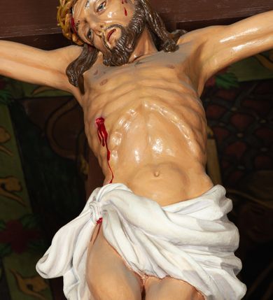 Zdjęcie nr 5: Trójfigurowa grupa Ukrzyżowania, rzeźby pełne. W środku znajduje się krucyfiks z postacią umarłego Chrystusa przybitego trzema gwoździami do krzyża. Chrystus w lekkim zwisie, o szeroko rozłożonych ramionach, bezwładny, głowa opuszczona na prawe ramię. Twarz szczupła, owalna, o prostym nosie, dużych oczach i na wpół otwartych ustach, okolona zarostem. Włosy w postaci delikatnych fal opadają na plecy i prawe ramię. Na głowie korona cierniowa. Ciało o wyraźnie zarysowanej anatomii. Perizonium w formie tkaniny przewiązanej na boku i odsłaniającej prawe biodro, z dłuższym brzegiem zwisającym po prawej stronie. Draperia gęsto fałdowana, o miękkich załamaniach materiału. Na szczycie pionowej belki titulus w formie banderoli z napisem „IN/RI”. Figura Matki Boskiej ustawiona po lewej stronie, zwrócona lekko w stronę krzyża, z głową uniesioną w kierunku Chrystusa i uniesionymi rękami. Twarz podłużna, niemal bez cech indywidualnych. Ubrana w długą białą suknię ze złotym paskiem, zielony płaszcz ze złotym podbiciem okrywający głowę, ramiona i plecy. Figura Marii Magdaleny po prawej stronie. Święta klęczy odwrócona od krzyża, z uniesioną głową i rozłożonymi rękami. Twarz młoda, owalna o słabo zindywidualizowanych rysach, włosy w postaci loków opadają na plecy. Jest ubrana w różową suknię ze złotym pasem oraz niebieski płaszcz ze złotym podbiciem. Polichromia w partiach ciała naturalistyczna. 