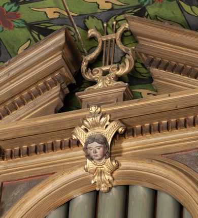 Zdjęcie nr 3: Organy architektoniczne, trójosiowe, na podwójnym cokole. Osie wydzielone pilastrami dźwigającymi gzyms z kostkowaniem. Środkowa wyższa niż boczne, zwieńczona trójkątnym, przerwanym przyczółkiem; pośrodku na gzymsie, rzeźbiona lira na cokole. Na osiach prześwity na piszczałki w formie stojącego prostokąta zamkniętego łukiem półkolistym, flankowanym przez półpilastry podtrzymujące profilowaną archiwoltę zwieńczoną kluczem; w osiach bocznych klucz w postaci liścia, w środkowej kartusz z głową. Zewnętrzne pilastry dekorowane kaboszonami i kampanulami, na gzymsie osi bocznych wazony. Struktura bejcowana w kolorze brązowym, ornamentyka złocona.