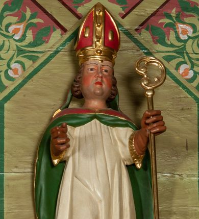 Zdjęcie nr 1: Figura ścięta, na cokole. Święty Stanisław został ukazany w pozycji stojącej z uniesionymi rękami, w lewej dłoni trzymając pastorał. Twarz ma pociągłą, o drobnych rysach. Jest ubrany w fioletową albę, białą komżę  z pasem złotej koronki u spodu oraz czerwoną kapę podbitą zielenią. Włosy ma kręcone, zaczesane do tyłu, na głowie czerwoną infułę ze złotymi ozdobami; rękawiczki i buty brązowe. Po prawej stronie, u stóp biskupa ukazany został Piotrowin, w popiersiu, z głową wzniesioną w stronę św. Stanisława, z rękami złożonymi w geście modlitewnym. Ma bujny zarost, jest ubrany w szarą koszulę. Polichromia w odsłoniętych partiach ciała naturalistyczna.