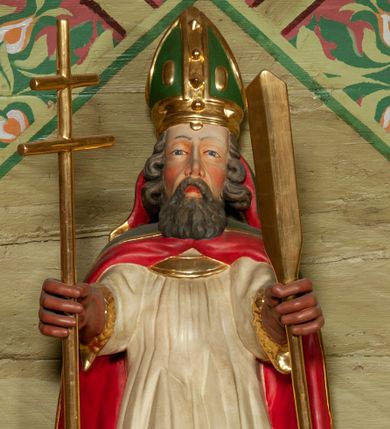 Zdjęcie nr 1: Figura ścięta, na cokole. Święty Wojciech przedstawiony w całej postaci, stojący frontalnie, obie ręce ma zgięte w łokciach, w prawej trzyma ferulę, czyli pastorał zakończony podwójnym krzyżem, w lewej wiosło. Wojciech ma pociągła twarz z dużymi, szarymi oczami, wąskim nosem i ustami o kącikach skierowanych w dół. Ciemne, siwiejące włosy ma zaczesane do tyłu, gęsta, długa broda pasmami opada na pierś. Jest ubrany w strój biskupi: fioletową albę i białą rokietę zdobioną złotym pasem koronki przy dolnej krawędzi. Na to założoną ma obszerną, czerwoną kapę, która spięta pod szyją okrywa ramiona i spływa do ziemi; na głowie ma infułę, rękawiczki i buty brązowe. Polichromia w odkrytych partiach karnacji postaci traktowana naturalistycznie.