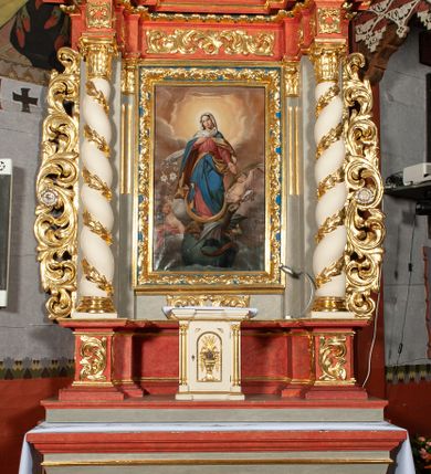 Zdjęcie nr 4: Obraz w kształcie stojącego prostokąta ujętego profilowaną ramą z motywem złoconej dekoracji roślinnej na błękitnym tle. Maria w ujęciu całopostaciowym, w kontrapoście. Głowa lekko przechylona w prawo, okryta długą białą chustą, przechodzącą na prawe ramię. Twarz o delikatnych, młodzieńczych rysach, lekkim rumieńcu. Wzrok skierowany ku górze. Prawa ręka złożona na piersiach, lewa wyprostowana, opuszczona w dół. Maria ubrana w długą, jasnoczerwoną tunikę, przewiązaną czerwonym paskiem oraz niebieski płaszcz z żółtym podszyciem, wywinięty wokół lewej ręki. Wokół głowy jasny, świetlisty nimb. Pod prawą stopą sierp księżyca, a pod nim uskrzydlony smok, trzymający w szponach jabłko, siedzący na globie ziemskim. Szarobrązowy stwór na wyszczerzone kły i rozłożone skrzydła. Po bokach Marii para nagich aniołków, z których umieszczony po lewej trzyma kwiat lilii, a po prawej gałązkę palmową. Scena ukazana na tle obłoków. Kolorystyka nasycona, o ciepłej gamie barwnej.