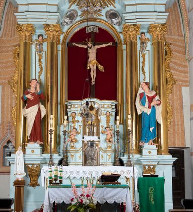 Zdjęcie nr 2: Nastawa architektoniczna, jednoosiowa, jednokondygnacyjna, ustawiona na wysokim cokole. 
Ołtarz drewniany, prostopadłościenny. Na nim tabernakulum z tronem wystawienia. Tabernakulum w formie drewnianej, prostopadłościennej skrzynki, obitej blachą, o wypukłej ściance frontowej, zamkniętej profilowanym gzymsem. Drzwiczki w formie stojącego prostokąta zamkniętego łukiem półkolistym, obwiedzione ramą, z wizerunkiem baranka z krzyżem, leżącego na księdze z pieczęciami, w glorii. Tabernakulum w drewnianej obudowie, wpisane w wysoki cokół, na nim tron wystawienia w formie obrotowej niszy w kształcie stojącego prostokąta, z podwieszoną draperią. Nisza flankowana parą pilastrów, przechodzących w spływy wolutowe, na których siedzą putta. Pilastry dźwigają pełne belkowanie, wyłamane na ich osi, częściowo przerwane przez niszę. Pilastry dekorowane uskrzydlonymi główkami anielskimi oraz snopkiem zboża i kiściami winogron. Cały tron wystawienia dekorowany rocaille'm. 
Nastawa ujęta dwiema parami kolumn o kapitelach kompozytowych, dźwigających pełne belkowanie, wyłamane pomiędzy nimi, przerwane nad polem środkowym. Na belkowaniu fragmenty przerwanego, półkolistego przyczółka, na których leżą putta. Całe pole środkowe wypełnia wnęka w formie stojącego prostokąta zamkniętego łukiem półkolistym, ujęta profilowaną ramą, z figurą Chrystusa Ukrzyżowanego. Nad łukiem wnęki, pomiędzy obłokami, gołębica Ducha Świętego w glorii promienistej. Pomiędzy kolumnami płyciny z uskrzydlonymi główkami anielskimi na obłokach, rocaille oraz figury św.  Jana Ewangelisty i Matki Boskiej na rocaille'owych wspornikach. Kondygnacja ujęta uszami utworzonymi z rocaille'u. Nastawa zwieńczona rzeźbą tronującego Boga Ojca z globem i berłem, pośród obłoków i uskrzydlonych główek anielskich, w glorii promienistej. 
Struktura polichromowana w kolorze błękitnym, kolumny, profile i ornamentyka złocone. 
