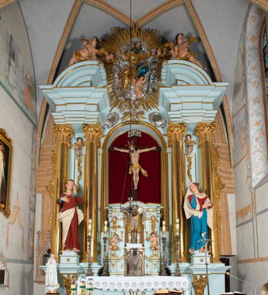 Zdjęcie nr 1: Nastawa architektoniczna, jednoosiowa, jednokondygnacyjna, ustawiona na wysokim cokole. 
Ołtarz drewniany, prostopadłościenny. Na nim tabernakulum z tronem wystawienia. Tabernakulum w formie drewnianej, prostopadłościennej skrzynki, obitej blachą, o wypukłej ściance frontowej, zamkniętej profilowanym gzymsem. Drzwiczki w formie stojącego prostokąta zamkniętego łukiem półkolistym, obwiedzione ramą, z wizerunkiem baranka z krzyżem, leżącego na księdze z pieczęciami, w glorii. Tabernakulum w drewnianej obudowie, wpisane w wysoki cokół, na nim tron wystawienia w formie obrotowej niszy w kształcie stojącego prostokąta, z podwieszoną draperią. Nisza flankowana parą pilastrów, przechodzących w spływy wolutowe, na których siedzą putta. Pilastry dźwigają pełne belkowanie, wyłamane na ich osi, częściowo przerwane przez niszę. Pilastry dekorowane uskrzydlonymi główkami anielskimi oraz snopkiem zboża i kiściami winogron. Cały tron wystawienia dekorowany rocaille&#039;m. 
Nastawa ujęta dwiema parami kolumn o kapitelach kompozytowych, dźwigających pełne belkowanie, wyłamane pomiędzy nimi, przerwane nad polem środkowym. Na belkowaniu fragmenty przerwanego, półkolistego przyczółka, na których leżą putta. Całe pole środkowe wypełnia wnęka w formie stojącego prostokąta zamkniętego łukiem półkolistym, ujęta profilowaną ramą, z figurą Chrystusa Ukrzyżowanego. Nad łukiem wnęki, pomiędzy obłokami, gołębica Ducha Świętego w glorii promienistej. Pomiędzy kolumnami płyciny z uskrzydlonymi główkami anielskimi na obłokach, rocaille oraz figury św.  Jana Ewangelisty i Matki Boskiej na rocaille&#039;owych wspornikach. Kondygnacja ujęta uszami utworzonymi z rocaille&#039;u. Nastawa zwieńczona rzeźbą tronującego Boga Ojca z globem i berłem, pośród obłoków i uskrzydlonych główek anielskich, w glorii promienistej. 
Struktura polichromowana w kolorze błękitnym, kolumny, profile i ornamentyka złocone. 
