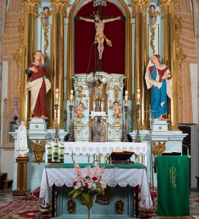 Zdjęcie nr 24: Nastawa architektoniczna, jednoosiowa, jednokondygnacyjna, ustawiona na wysokim cokole. 
Ołtarz drewniany, prostopadłościenny. Na nim tabernakulum z tronem wystawienia. Tabernakulum w formie drewnianej, prostopadłościennej skrzynki, obitej blachą, o wypukłej ściance frontowej, zamkniętej profilowanym gzymsem. Drzwiczki w formie stojącego prostokąta zamkniętego łukiem półkolistym, obwiedzione ramą, z wizerunkiem baranka z krzyżem, leżącego na księdze z pieczęciami, w glorii. Tabernakulum w drewnianej obudowie, wpisane w wysoki cokół, na nim tron wystawienia w formie obrotowej niszy w kształcie stojącego prostokąta, z podwieszoną draperią. Nisza flankowana parą pilastrów, przechodzących w spływy wolutowe, na których siedzą putta. Pilastry dźwigają pełne belkowanie, wyłamane na ich osi, częściowo przerwane przez niszę. Pilastry dekorowane uskrzydlonymi główkami anielskimi oraz snopkiem zboża i kiściami winogron. Cały tron wystawienia dekorowany rocaille'm. 
Nastawa ujęta dwiema parami kolumn o kapitelach kompozytowych, dźwigających pełne belkowanie, wyłamane pomiędzy nimi, przerwane nad polem środkowym. Na belkowaniu fragmenty przerwanego, półkolistego przyczółka, na których leżą putta. Całe pole środkowe wypełnia wnęka w formie stojącego prostokąta zamkniętego łukiem półkolistym, ujęta profilowaną ramą, z figurą Chrystusa Ukrzyżowanego. Nad łukiem wnęki, pomiędzy obłokami, gołębica Ducha Świętego w glorii promienistej. Pomiędzy kolumnami płyciny z uskrzydlonymi główkami anielskimi na obłokach, rocaille oraz figury św.  Jana Ewangelisty i Matki Boskiej na rocaille'owych wspornikach. Kondygnacja ujęta uszami utworzonymi z rocaille'u. Nastawa zwieńczona rzeźbą tronującego Boga Ojca z globem i berłem, pośród obłoków i uskrzydlonych główek anielskich, w glorii promienistej. 
Struktura polichromowana w kolorze błękitnym, kolumny, profile i ornamentyka złocone. 
