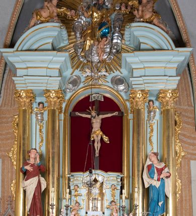 Zdjęcie nr 13: Nastawa architektoniczna, jednoosiowa, jednokondygnacyjna, ustawiona na wysokim cokole. 
Ołtarz drewniany, prostopadłościenny. Na nim tabernakulum z tronem wystawienia. Tabernakulum w formie drewnianej, prostopadłościennej skrzynki, obitej blachą, o wypukłej ściance frontowej, zamkniętej profilowanym gzymsem. Drzwiczki w formie stojącego prostokąta zamkniętego łukiem półkolistym, obwiedzione ramą, z wizerunkiem baranka z krzyżem, leżącego na księdze z pieczęciami, w glorii. Tabernakulum w drewnianej obudowie, wpisane w wysoki cokół, na nim tron wystawienia w formie obrotowej niszy w kształcie stojącego prostokąta, z podwieszoną draperią. Nisza flankowana parą pilastrów, przechodzących w spływy wolutowe, na których siedzą putta. Pilastry dźwigają pełne belkowanie, wyłamane na ich osi, częściowo przerwane przez niszę. Pilastry dekorowane uskrzydlonymi główkami anielskimi oraz snopkiem zboża i kiściami winogron. Cały tron wystawienia dekorowany rocaille'm. 
Nastawa ujęta dwiema parami kolumn o kapitelach kompozytowych, dźwigających pełne belkowanie, wyłamane pomiędzy nimi, przerwane nad polem środkowym. Na belkowaniu fragmenty przerwanego, półkolistego przyczółka, na których leżą putta. Całe pole środkowe wypełnia wnęka w formie stojącego prostokąta zamkniętego łukiem półkolistym, ujęta profilowaną ramą, z figurą Chrystusa Ukrzyżowanego. Nad łukiem wnęki, pomiędzy obłokami, gołębica Ducha Świętego w glorii promienistej. Pomiędzy kolumnami płyciny z uskrzydlonymi główkami anielskimi na obłokach, rocaille oraz figury św.  Jana Ewangelisty i Matki Boskiej na rocaille'owych wspornikach. Kondygnacja ujęta uszami utworzonymi z rocaille'u. Nastawa zwieńczona rzeźbą tronującego Boga Ojca z globem i berłem, pośród obłoków i uskrzydlonych główek anielskich, w glorii promienistej. 
Struktura polichromowana w kolorze błękitnym, kolumny, profile i ornamentyka złocone. 
