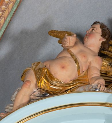 Zdjęcie nr 42: Nastawa architektoniczna, jednoosiowa, jednokondygnacyjna, ustawiona na wysokim cokole. 
Ołtarz drewniany, prostopadłościenny. Na nim tabernakulum z tronem wystawienia. Tabernakulum w formie drewnianej, prostopadłościennej skrzynki, obitej blachą, o wypukłej ściance frontowej, zamkniętej profilowanym gzymsem. Drzwiczki w formie stojącego prostokąta zamkniętego łukiem półkolistym, obwiedzione ramą, z wizerunkiem baranka z krzyżem, leżącego na księdze z pieczęciami, w glorii. Tabernakulum w drewnianej obudowie, wpisane w wysoki cokół, na nim tron wystawienia w formie obrotowej niszy w kształcie stojącego prostokąta, z podwieszoną draperią. Nisza flankowana parą pilastrów, przechodzących w spływy wolutowe, na których siedzą putta. Pilastry dźwigają pełne belkowanie, wyłamane na ich osi, częściowo przerwane przez niszę. Pilastry dekorowane uskrzydlonymi główkami anielskimi oraz snopkiem zboża i kiściami winogron. Cały tron wystawienia dekorowany rocaille&#039;m. 
Nastawa ujęta dwiema parami kolumn o kapitelach kompozytowych, dźwigających pełne belkowanie, wyłamane pomiędzy nimi, przerwane nad polem środkowym. Na belkowaniu fragmenty przerwanego, półkolistego przyczółka, na których leżą putta. Całe pole środkowe wypełnia wnęka w formie stojącego prostokąta zamkniętego łukiem półkolistym, ujęta profilowaną ramą, z figurą Chrystusa Ukrzyżowanego. Nad łukiem wnęki, pomiędzy obłokami, gołębica Ducha Świętego w glorii promienistej. Pomiędzy kolumnami płyciny z uskrzydlonymi główkami anielskimi na obłokach, rocaille oraz figury św.  Jana Ewangelisty i Matki Boskiej na rocaille&#039;owych wspornikach. Kondygnacja ujęta uszami utworzonymi z rocaille&#039;u. Nastawa zwieńczona rzeźbą tronującego Boga Ojca z globem i berłem, pośród obłoków i uskrzydlonych główek anielskich, w glorii promienistej. 
Struktura polichromowana w kolorze błękitnym, kolumny, profile i ornamentyka złocone. 
