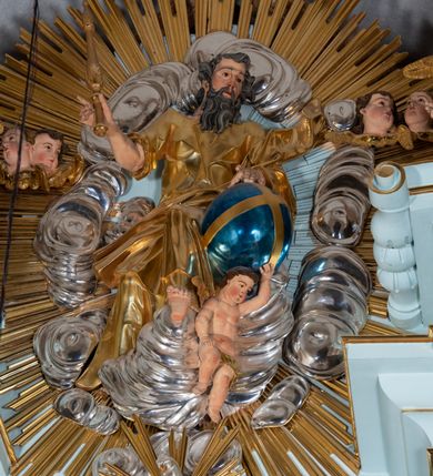 Zdjęcie nr 50: Nastawa architektoniczna, jednoosiowa, jednokondygnacyjna, ustawiona na wysokim cokole. 
Ołtarz drewniany, prostopadłościenny. Na nim tabernakulum z tronem wystawienia. Tabernakulum w formie drewnianej, prostopadłościennej skrzynki, obitej blachą, o wypukłej ściance frontowej, zamkniętej profilowanym gzymsem. Drzwiczki w formie stojącego prostokąta zamkniętego łukiem półkolistym, obwiedzione ramą, z wizerunkiem baranka z krzyżem, leżącego na księdze z pieczęciami, w glorii. Tabernakulum w drewnianej obudowie, wpisane w wysoki cokół, na nim tron wystawienia w formie obrotowej niszy w kształcie stojącego prostokąta, z podwieszoną draperią. Nisza flankowana parą pilastrów, przechodzących w spływy wolutowe, na których siedzą putta. Pilastry dźwigają pełne belkowanie, wyłamane na ich osi, częściowo przerwane przez niszę. Pilastry dekorowane uskrzydlonymi główkami anielskimi oraz snopkiem zboża i kiściami winogron. Cały tron wystawienia dekorowany rocaille&#039;m. 
Nastawa ujęta dwiema parami kolumn o kapitelach kompozytowych, dźwigających pełne belkowanie, wyłamane pomiędzy nimi, przerwane nad polem środkowym. Na belkowaniu fragmenty przerwanego, półkolistego przyczółka, na których leżą putta. Całe pole środkowe wypełnia wnęka w formie stojącego prostokąta zamkniętego łukiem półkolistym, ujęta profilowaną ramą, z figurą Chrystusa Ukrzyżowanego. Nad łukiem wnęki, pomiędzy obłokami, gołębica Ducha Świętego w glorii promienistej. Pomiędzy kolumnami płyciny z uskrzydlonymi główkami anielskimi na obłokach, rocaille oraz figury św.  Jana Ewangelisty i Matki Boskiej na rocaille&#039;owych wspornikach. Kondygnacja ujęta uszami utworzonymi z rocaille&#039;u. Nastawa zwieńczona rzeźbą tronującego Boga Ojca z globem i berłem, pośród obłoków i uskrzydlonych główek anielskich, w glorii promienistej. 
Struktura polichromowana w kolorze błękitnym, kolumny, profile i ornamentyka złocone. 
